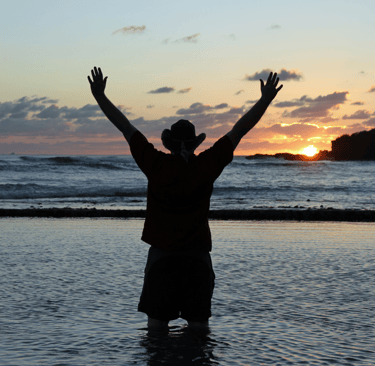 Un homme avec un chapeau qui se tiens devant un coucher du soleil a la plage 