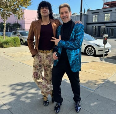 Tim and Bollywood actor in fashionable blazers posing on a sunny urban sidewalk with city buildings.
