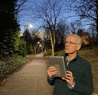 Photograph of a man, standing at the side of a footpath, at night, holding a tablet computer.