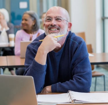 Mature man learner in a class wearing glasses smiling with laptop and mature women background