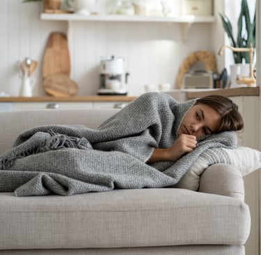 Frau liegt eingehüllt in eine graue Decke auf einem Sofa und ruht sich aus, im Hintergrund eine gemü