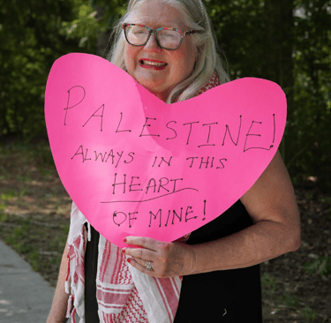 a woman wearing a keffiyeh stands holding a sign that says "palestine! always in this heart of mine"