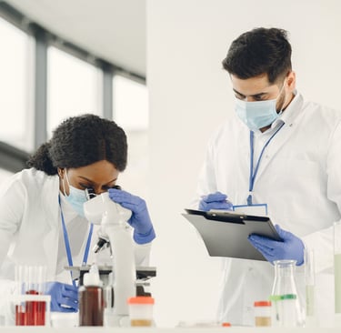 Woman in lab coat sitting looks into  a microscope while a man in lab coat stands with chart holder
