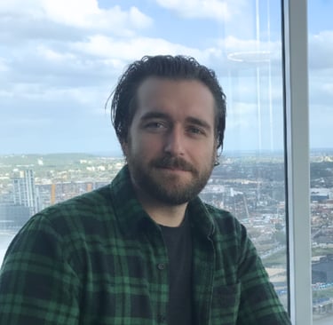 Professional man in a green flannel shirt posing by a window with a city skyline view.