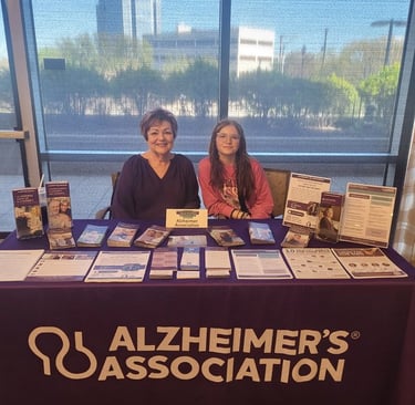 Photo of Debbie & colleague seated at an Alzheimer's Association info table, in front of a shaded window and urban landscape.