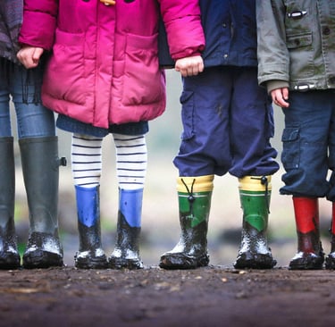 Four children standing in a row wearing colorful winter coats and muddy rain boots outdoors.
