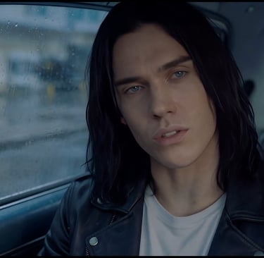 A man with long dark hair and blue eyes wearing a black leather jacket inside a car on a rainy day.