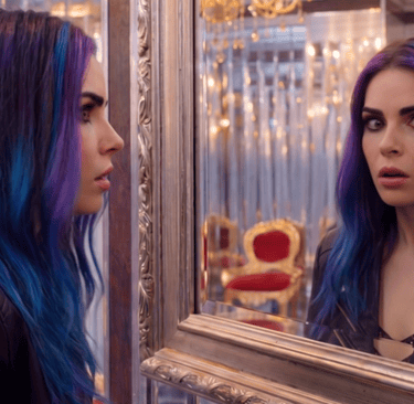 A woman with vibrant purple and blue hair looks surprised while staring at her reflection in a luxury mirror.