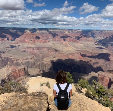 A hiker with a backpack sits on a rocky cliff overlooking the vast Grand Canyon landscape under a cloudy blue sky.