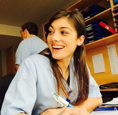 A smiling young female nurse in light blue medical scrubs writing notes at a hospital workstation.