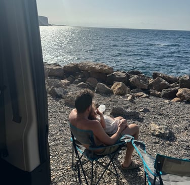 Father feeding a baby on a rocky beach while van life camping by the Mediterranean Sea.