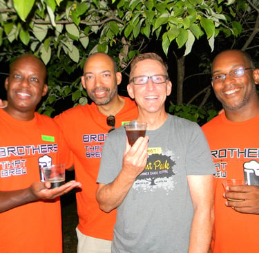 Four men smiling, wearing Brothers That Brew shirts and holding small tasting glasses.