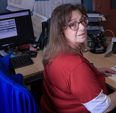 A female office professional wearing glasses and a red shirt reviews documents at her computer desk.