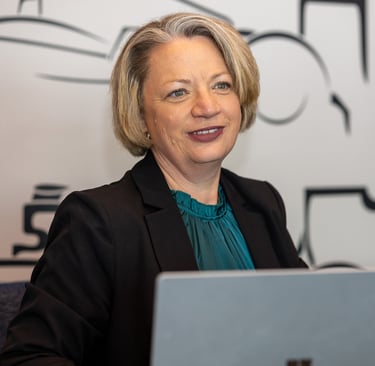 A smiling woman with short blonde hair and glasses, wearing a dark blazer over a teal top, sits at a desk with a laptop.