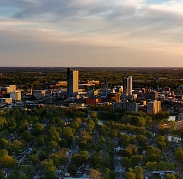 Aerial view of Fort Wayne Indiana skyline at sunset with St. Joseph River and lush green trees.