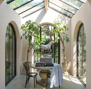 Sunlit garden room with a glass roof, arched windows, indoor trees, and a stone table with a horse sculpture.