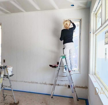 A woman on a ladder paints white textured wall panels during a modern home interior renovation project.