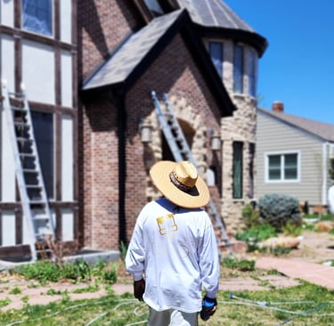 Professional house painter in a sun hat preparing to paint a brick Tudor home exterior with ladders.