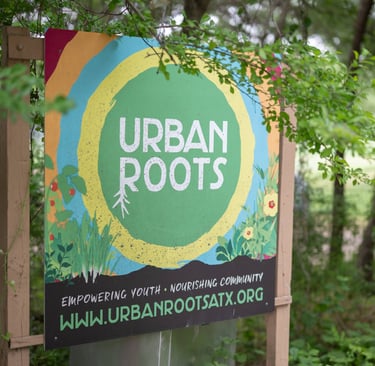 A colorful sign for Urban Roots stands nestled among lush greenery, partially framed by branches