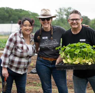 Three volunteers smile in a garden, holding fresh seedlings and tools, ready to plant and grow.