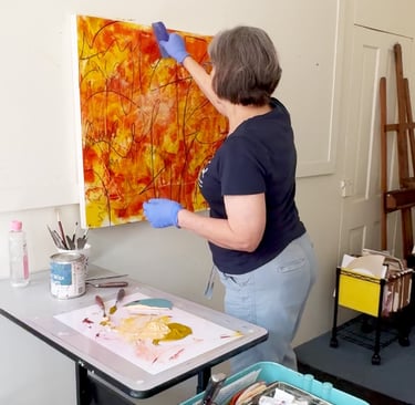 Artist in blue gloves applying cold wax medium to a large orange and yellow abstract painting.