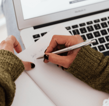 Hand holding a notebook page and writing with laptop in the background