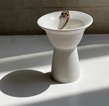 A wheel thrown, white porcelain bowl with an elegant pedestal base. On its rim is a cast bronze knot