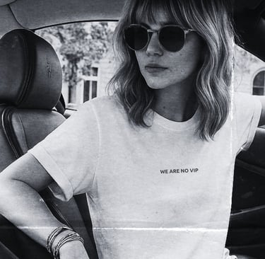 Black-and-white photo of a woman in sunglasses wearing the NOVIP ‘We Are No VIP’ T-shirt inside a ca