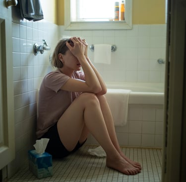A distressed woman sits on a bathroom floor holding her head, illustrating mental health struggles and stress.