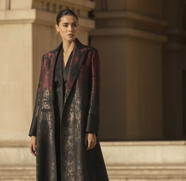 Elegant model wearing a luxury red and black brocade trench coat outdoors with classic architecture.