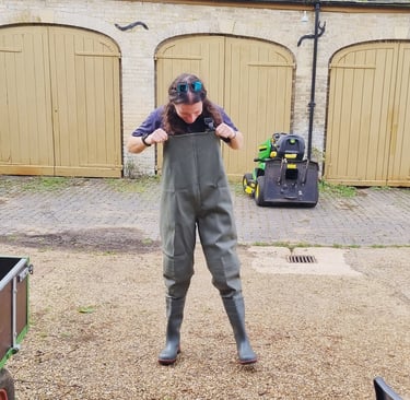 A woman putting on waterproof olive green fishing chest waders in a gravel courtyard.