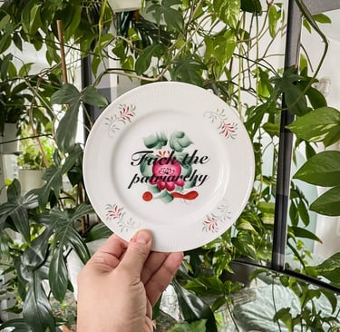 Hand holding a vintage decorative plate with floral patterns and Fuck the patriarchy text against green houseplants.