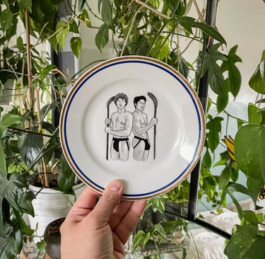A decorative ceramic plate featuring an illustration of two shirtless hockey players surrounded by green houseplants.