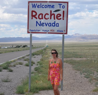 a woman standing in front of a welcome sign for Rachel, Nevada