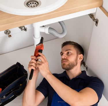 A professional residential plumber lies beneath a modern kitchen sink, repairing pipes.