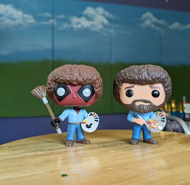Funko POP Deadpool Bob Ross and Bob Ross figurines standing in front of a 4-panel project painting in an art studio.