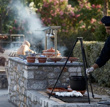 A professional chef cooking traditional Greek food over an outdoor stone grill with a hanging tripod pot.