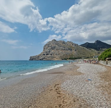 Sunny pebble beach in Rhodes with tourists swimming in turquoise water near a large rocky mountain.
