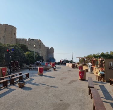 Medieval festival marketplace with wooden benches and shields near an ancient stone castle wall.