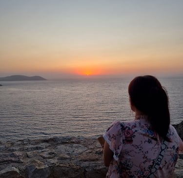 A woman in a floral shirt watches a peaceful ocean sunset from a rocky cliffside view.