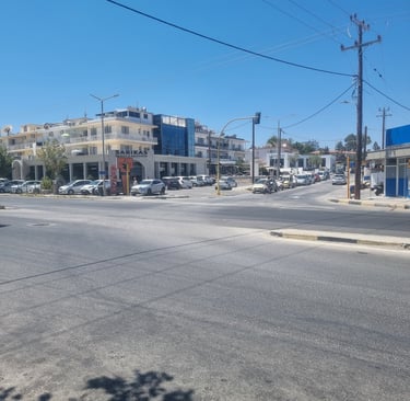 A sunny street intersection in Faliraki, featuring cars parked near shops and a clear blue sky.