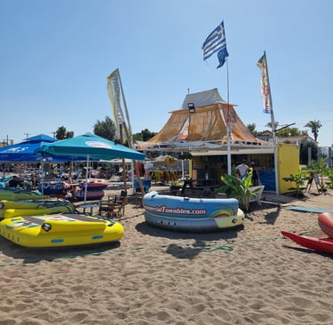 Inflatable water sports tubes and a beach hut bar on a sandy Greek shore under a blue sky.