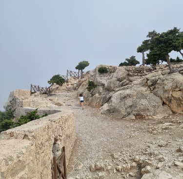 A hiker climbs a rocky mountain trail toward a medieval stone fortress under a cloudy sky.
