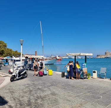 Tourists with colorful luggage at Mandraki Harbor in Rhodes Greece with sailboats and kiosks.