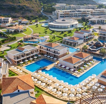 Aerial view of a luxury Greek resort with blue swimming pools, white umbrellas, and elegant villas.