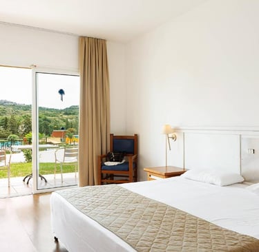 Bright modern hotel room with white bedding and a balcony overlooking a pool and hills.