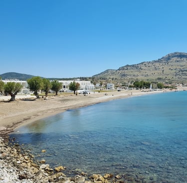 A sunny coastal beach scene with clear blue sea, a sandy shore, white buildings, and distant hills under a bright sky