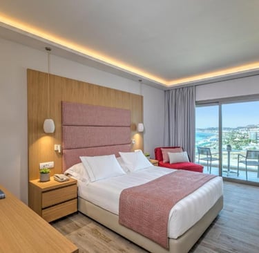 Modern hotel room with a double bed, pink headboard, and balcony overlooking the ocean.