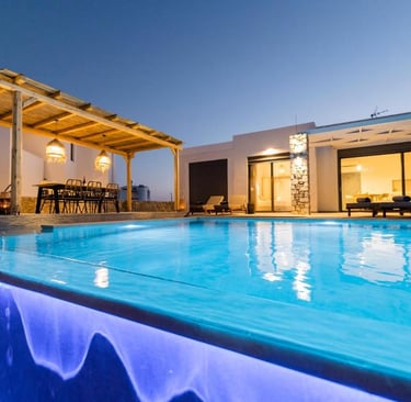Luxury villa with an illuminated infinity pool and outdoor dining pergola at sunset.
