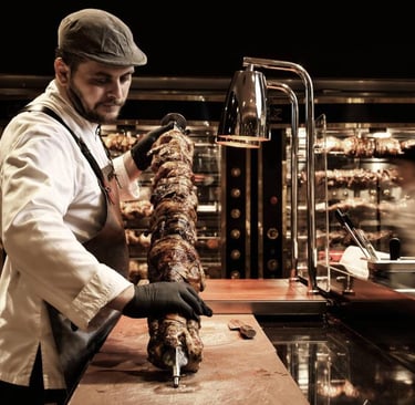 Professional chef preparing a vertical rotisserie spit of roasted meat in a restaurant kitchen.